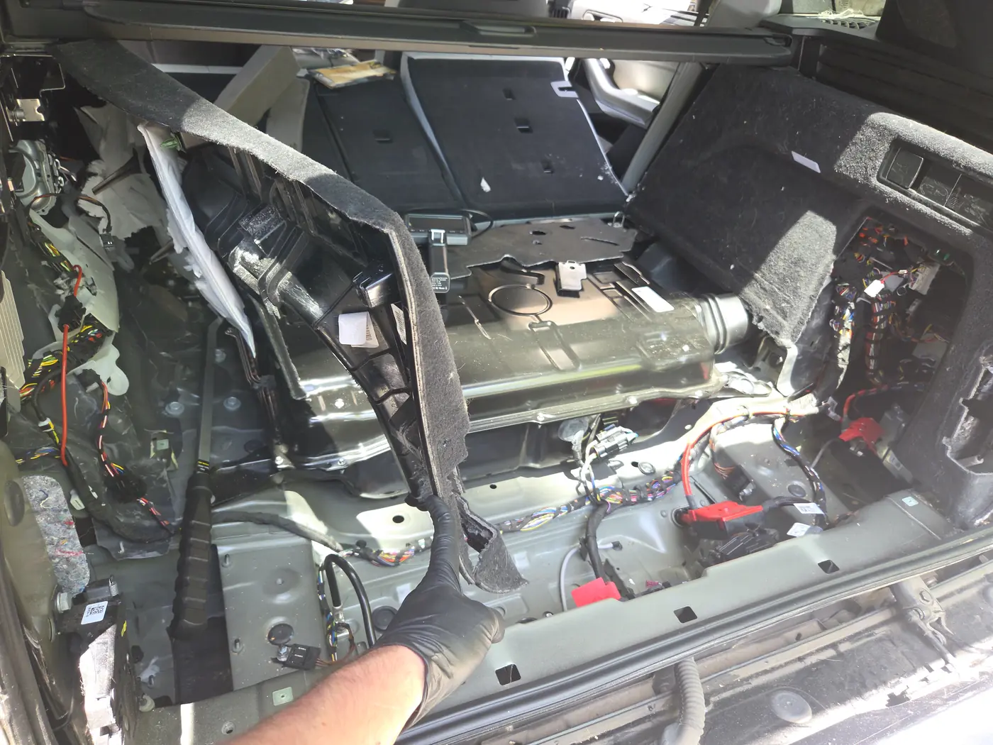 An SUV's cargo area stripped to bare metal with the trim pulled back and wiring exposed, a gloved hand holding a panel
