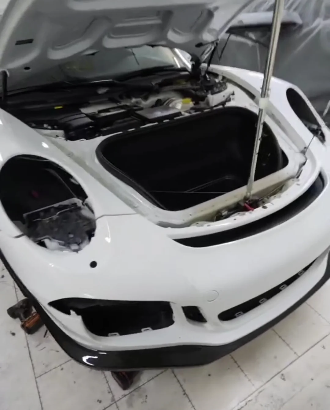 A white Porsche 911 with the front trunk open and the headlights out, on a garage floor