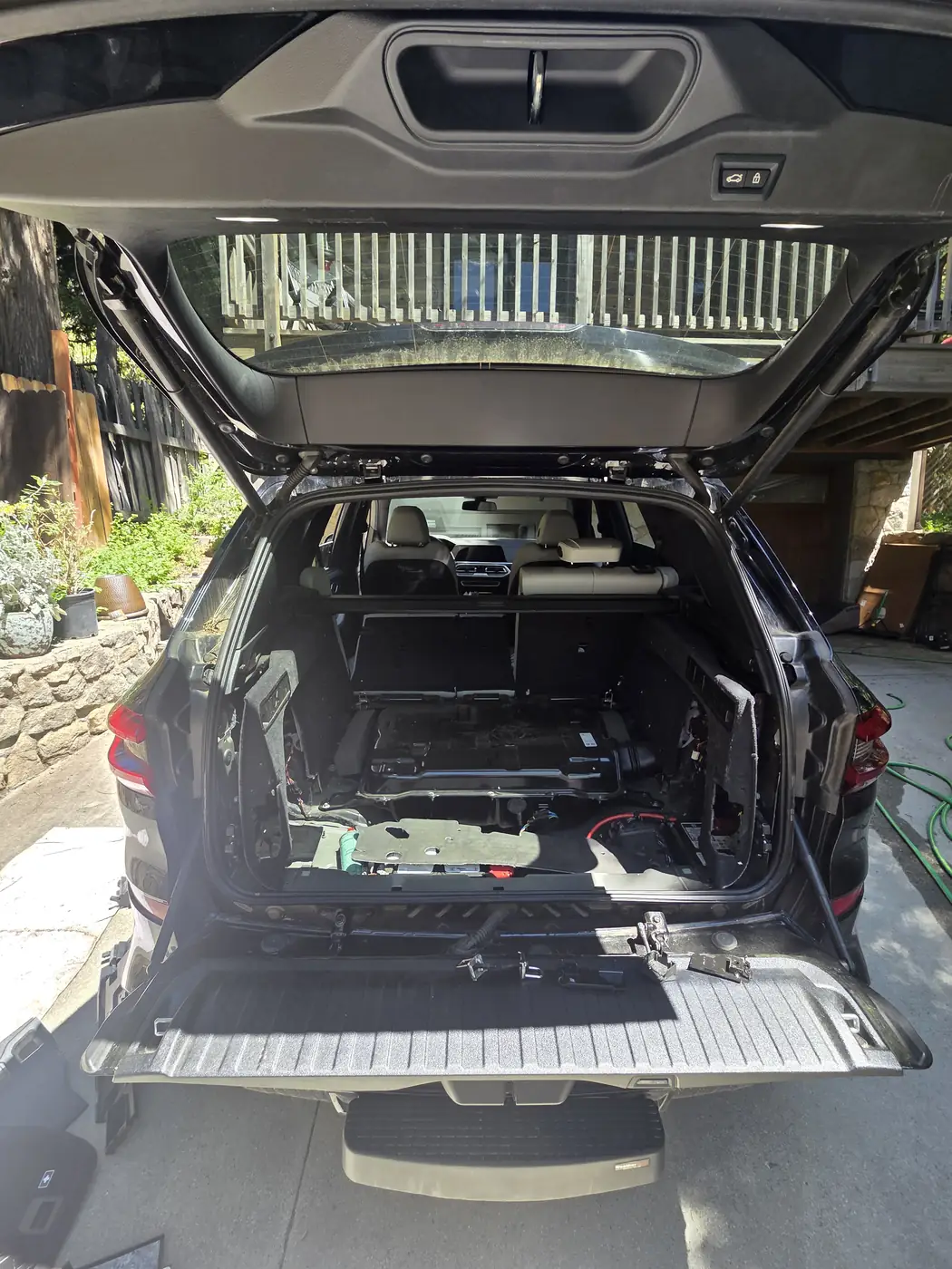 An SUV parked in a driveway with the tailgate open and the cargo floor out, plants and trees behind