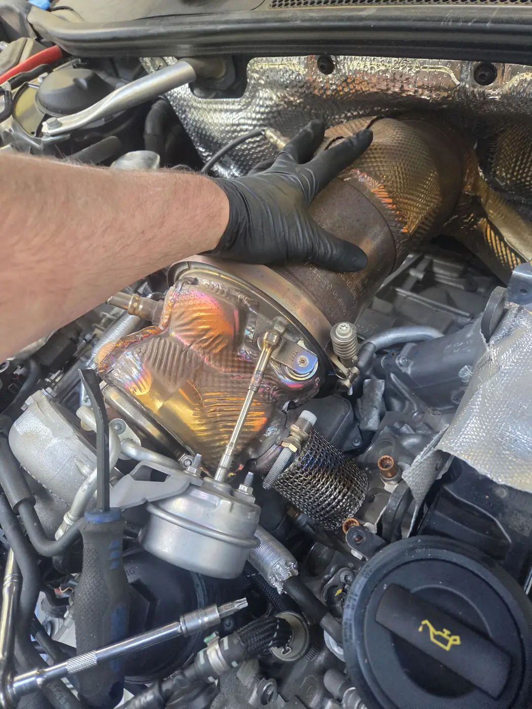 A gloved hand on a turbocharger and its heat shield deep in an engine bay